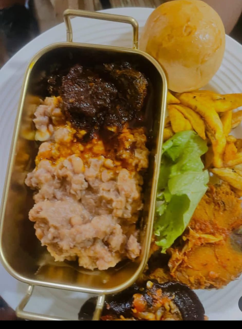 Beans porridge served in a gold dish with plantain, bread roll and fried fish